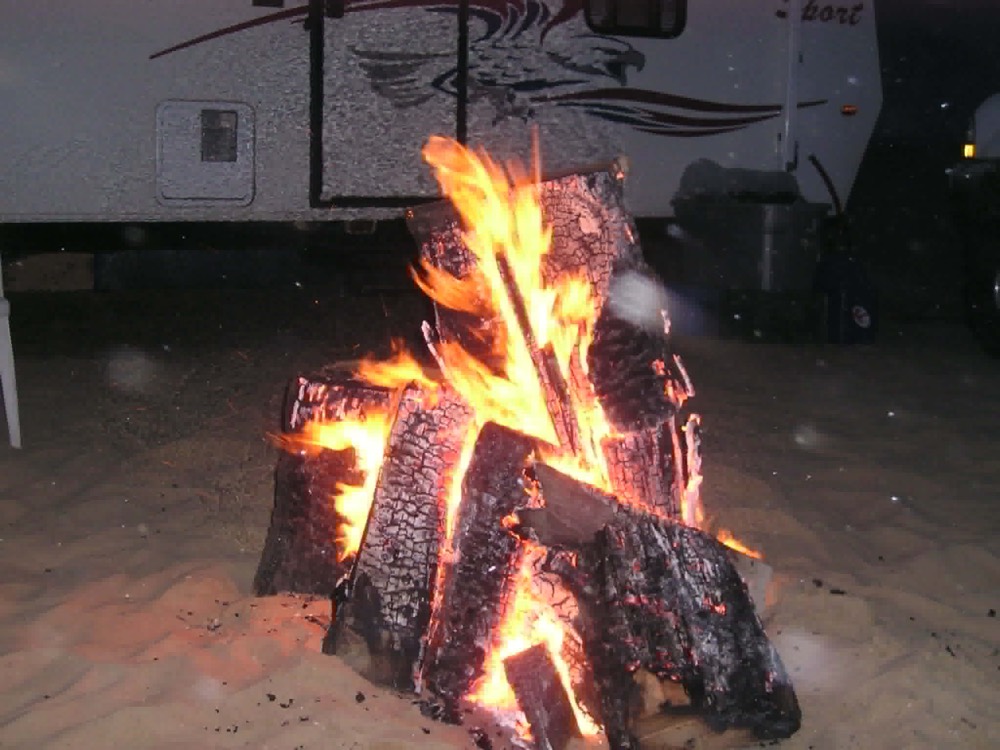 Driftwood campfire burning on the sand in front of a fifth-wheel trailer