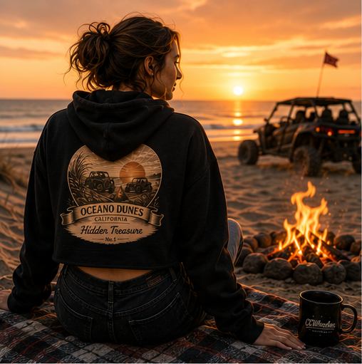 A rider in a black CCWheelers Sunset Collection crop hoodie sitting by a beach campfire at sunset with a UTV parked on the sand, Oceano Dunes California