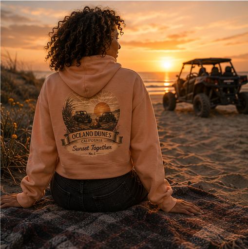A rider in a peach CCWheelers Sunset Collection crop hoodie sitting on the sand watching the sunset, Oceano Dunes California